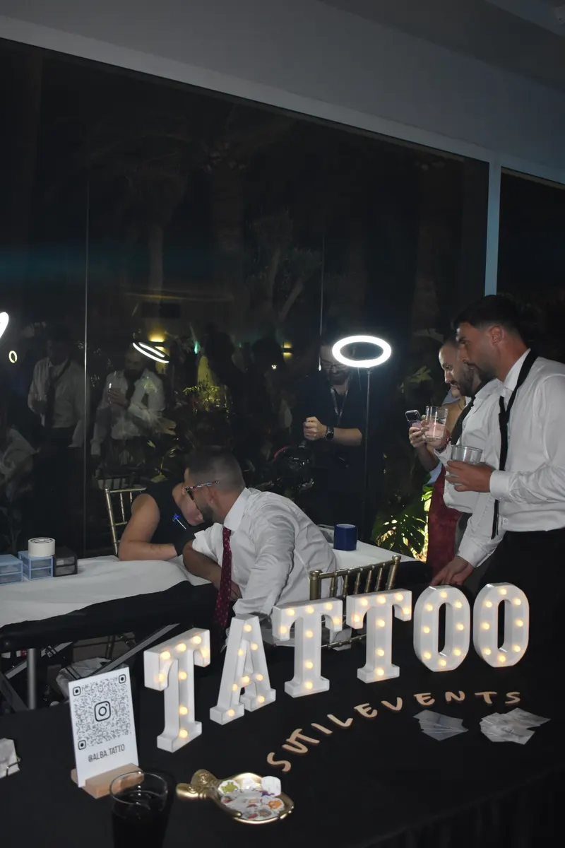 Rincón de tatuajes de SutilEvents de noche: letras luminosas TATTOO, letras de madera con el nombre y Alba tatuando a un invitado