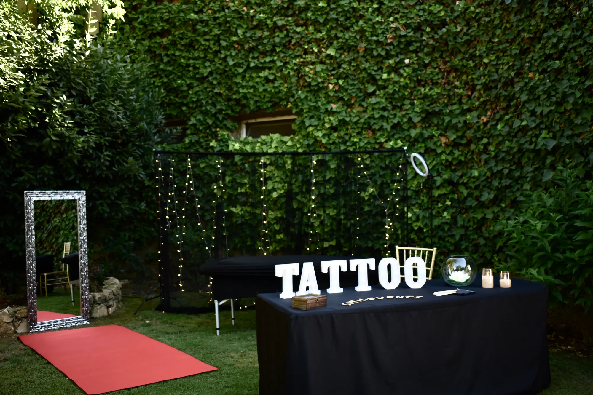 Rincón de mini tattoos montado en el jardín de una boda: pared de hiedra, cortina de luces, camilla negra, letras TATTOO, espejo y alfombra roja