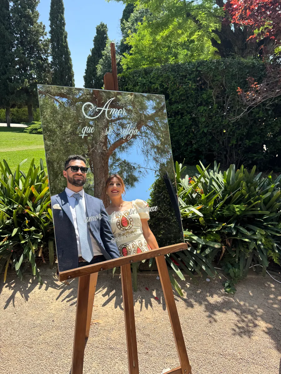 Pareja de novios posando detrás de un cartel de espejo con caligrafía blanca sobre un caballete de madera, en un jardín